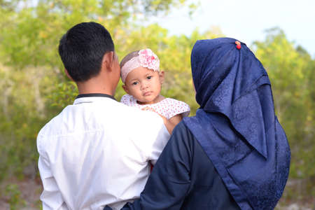 Midshot of a happy family outside on nature. Happy Young Asian Muslim mother and father hugs his daughterの写真素材