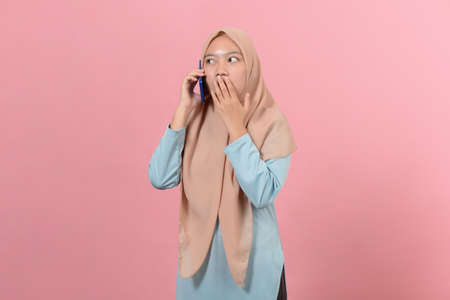 Young beautiful Asian muslim woman surprised shocked with mobile phone close mouth with palm, isolated on pink backgroundの写真素材