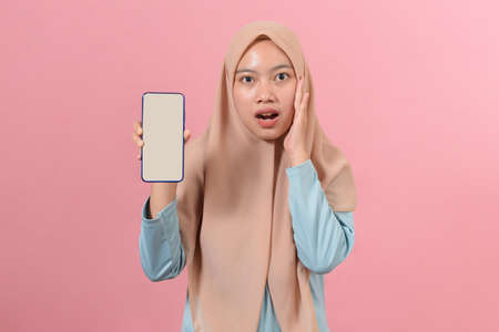 Shocked Asian Muslim Woman touching faces while showing smartphones with blank screen, against on pink backgroundの写真素材