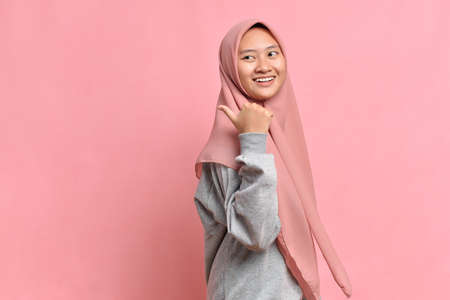 Asian muslim woman smiling and pointing to presenting something on her side, against pink background with copy spaceの写真素材