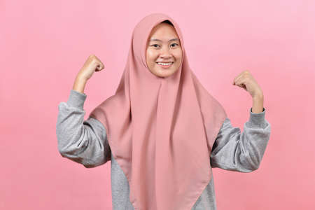 Studio shot of positive young Asian muslim woman raises arms shows muscles pretends to be very strong and powerful smiles gently wears hijab isolated over pink background.の写真素材