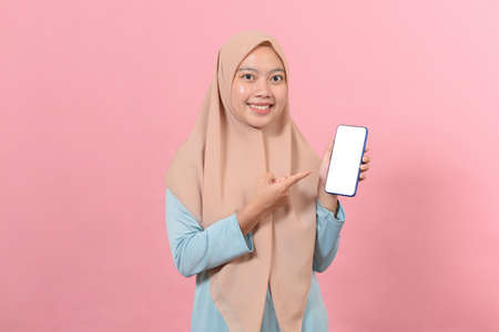 Portrait of a joyful asian muslim businesswoman showing blank screen mobile phone isolated over pink backgroundの写真素材