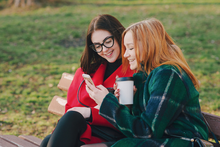 Girls walking around town on a sunny day with phones sitting on a bench and drink coffeeの写真素材