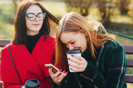 Girls walking around town on a sunny day with phones sitting on a bench and drink coffeeの写真素材