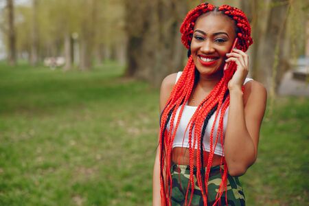 A young and stylish dark-skinned girl with red dreads walking in the summer park with phone