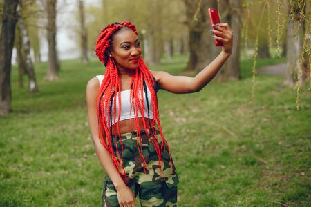 A young and stylish dark-skinned girl with red dreads walking in the summer park with phoneの写真素材