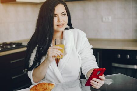 beautiful young girl with black hair and white robe sitting at home in the kitchen at the table and eating an croissant and drinking orange juiceの写真素材
