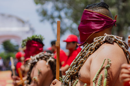 Man with covered face carries a wooden cross, wearing a crown of thorns and chains on his body during the Holy Week tradition known as "Los Engrillados" in Atlixco, Mexico.の写真素材