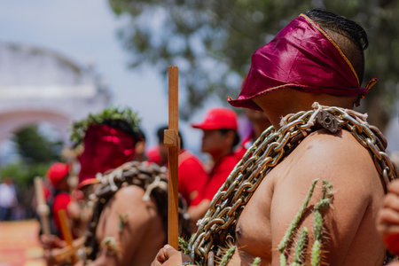 Man with covered face carries a wooden cross, wearing a crown of thorns and chains on his body during the Holy Week tradition known as "Los Engrillados" in Atlixco, Mexico.の写真素材