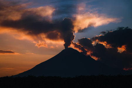 Powerful silhouette of Mexico's Popocatepetl Volcano during an ash exhalation, with a dramatic sky and twilight light rays at sunset.の写真素材