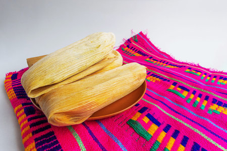 Angled view of several tamales wrapped in natural leaves on a clay plate and traditional pink tablecloth with embroidery.の写真素材