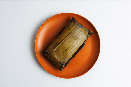 Top view of a steamed tamale in a banana leaf, served on an orange plate with a minimalist white...