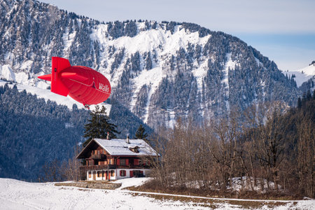 Chateau d'oex, Vaud Canton, Switzerland - 1st February 2026: Red Hot Air Balloon Wallis Airship Flying Over Alpine Chalet in Switzerland Winter.のeditorial素材