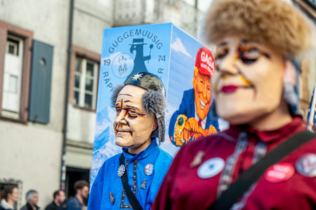 Basel, Switzerland - March 25, 2026: Masked carnival participants walking beside a satirical float...