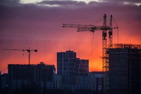 Silhouette of tower cranes at sunset. Urban landscape against a bright orange-purple sunsetの写真素材