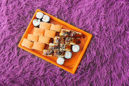 Set of different types of sushi rolls with shrimp, salmon, eel, in an orange box on a purple fluffy carpet or bedspread. Japanese Food Delivery Conceptの写真素材