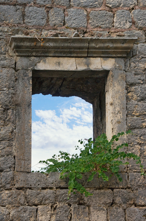 Wall of an old destroyed building with a window through which a blue sky with white clouds is visibleの写真素材
