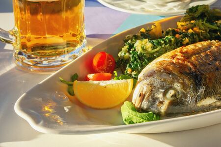 Balkan cuisine. Sunday lunch: mug of beer and grilled fish with green vegetables and slice of lemonの写真素材