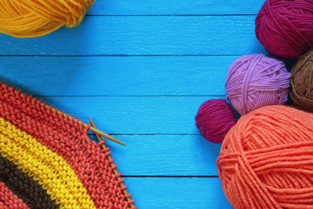 Hobby and knitting concepts.  Multi-colored clews  and balls of wool on blue rustic table. Unfinished striped knitwork with knitting needles. Copy spaceの写真素材