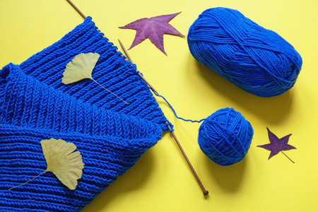 Autumn hobby concept, knitting. Bright warm blue scarf with knitting needles and ball of wool on yellow background. Autumn leavesの写真素材