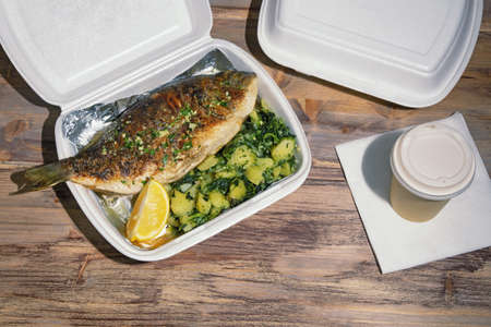 Food delivery. Grilled fish ( sea bream ) with green leafy vegetables and slice of lemon in takeout container on rustic table. Copy spaceの写真素材