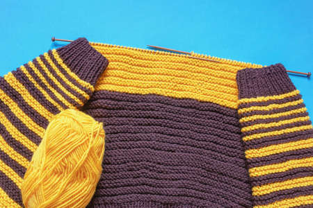 Home hobbies concept. Unfinished yellow and brown knitting, ball of wool and knitting  needles on blue background. Copy spaceの写真素材