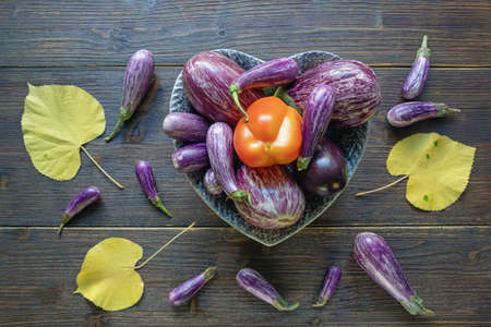 Autumn vegetables. Red bell pepper, different varieties of eggplant vegetables in heart shape plate and yellow leaves on dark rustic table. Healthy, vegetarian food concept. Flat lay, copy spaceの写真素材