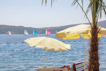 Beach vacation concepts.  Beach umbrellas on coast, colorful sailboats on water. Montenegro,  view of Kotor Bay near Tivat cityの写真素材