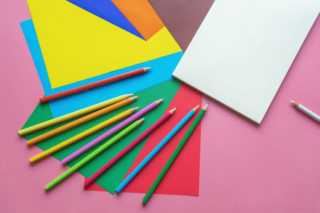 School and hobby concepts. Colored paper sheets, colored pencils and open sketchbook on pink paper background. Copy spaceの写真素材