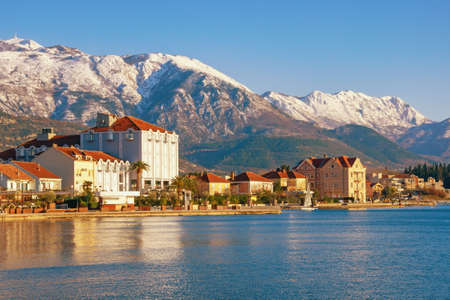Beautiful winter Mediterranean landscape. Green town against snow-capped mountains. View of embankment of Tivat city on sunny winter day. Montenegro, Adriatic Sea, Bay of Kotorの写真素材