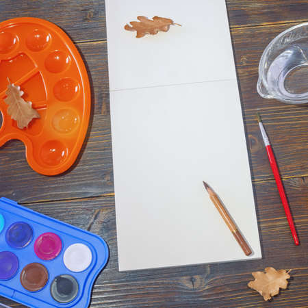 Sketchbook, watercolor paints, palette and dry autumn leaves on dark rustic table. School concept. Copy spaceの写真素材