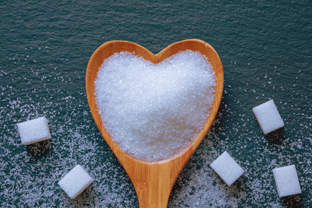 White crystalline sugar in heart-shaped spoon on black background. Lump sugar. Flat lay, copy space