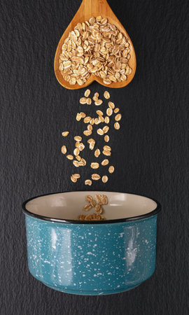 Cereal flakes in a heart-shaped wooden spoon fall into a small blue pan. Dark background,  copy...