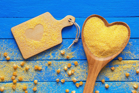 Cornmeal in heart-shaped wooden spoon. Cutting board with cornmeal, corn grains  on blue rustic background. Copy spaceの写真素材