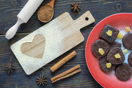 Cutting board with scattered flour in the shape of a heart. Delicious homemade cookies in a red plate. Ingrediends, dark rustic background, flat layの写真素材