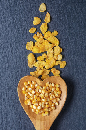Corn grains in a heart-shaped wooden spoon, corn flakes on a dark table. Flat lay, copy space