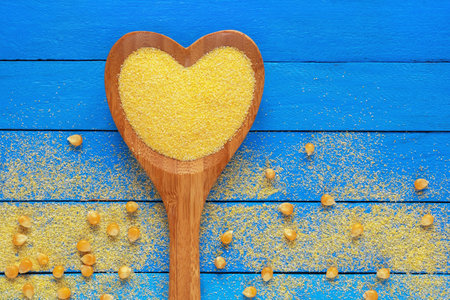 Cornmeal in heart-shaped spoon and corn grains on blue table. Copy spaceの写真素材
