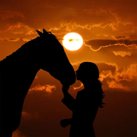 Silhouette of a young girl with horse giving him a kiss over a red sunset backgroundの写真素材