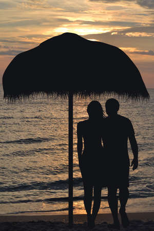 Romantic couple on the beach under straw umbrella hugging at sunriseの写真素材