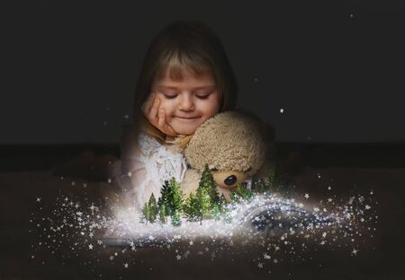 Little girl with her teddy bear, watching a enchanted forest  on the pages of an open magic bookの写真素材