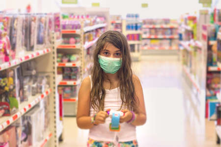 Little girl wearing disposable medical mask, while shopping in toys department, during coronavirus outbreakの写真素材