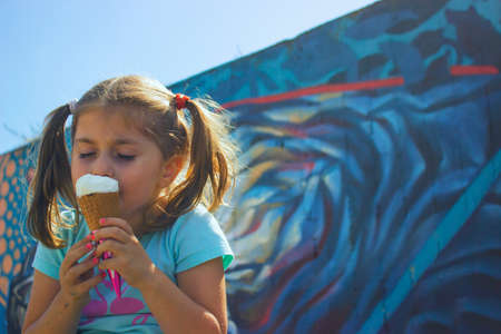 Little girl eating ice cream with her eyes closed, against blue wall