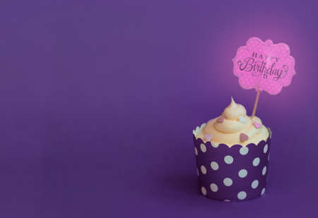 Vanilla cupcake with small decorative hearts and happy birthday sign, in violet baking paper cup, against violet backgroundの写真素材