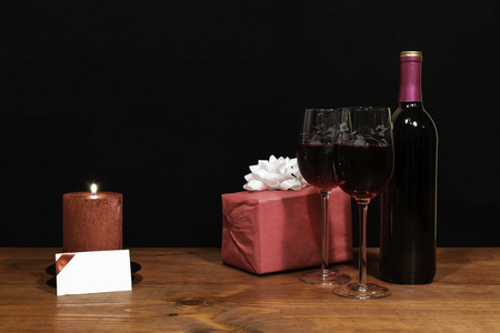 Beautiful etched wine glasses and bottle of red wine, red candle, wrapped present with bow on wooden table with name tag on dark background. Valentines, Mothers Day, Easter, Christmas, Wedding Conceptsの写真素材