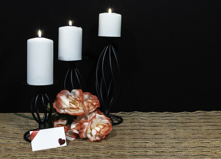 Beautiful orange and white roses, white candle perched on black candle holders on mesh place mat and wooden table with card and dark background. Valentines, Mothers Day, Easter, Christmas, Wedding Conceptsの写真素材
