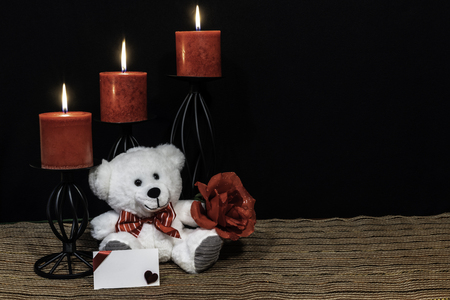 Cudlely teddy bear with red bow tie, red rose, red candles perched on black candle holders on mesh place mat and wooden table with card and dark background. Valentines, Mothers Day, Easter, Christmas, Wedding Conceptsの写真素材