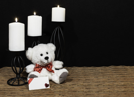 Cudlely teddy bear with red bow tie, white candles perched on black candle holders on mesh place mat and wooden table with card and dark background. Valentines, Mothers Day, Easter, Christmas, Wedding Conceptsの写真素材