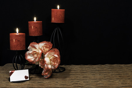 Beautiful orange and orange and white roses, red candle perched on black candle holders on mesh place mat and wooden table with dark background and card. Valentines, Mothers Day, Easter, Christmas, Wedding Conceptsの写真素材