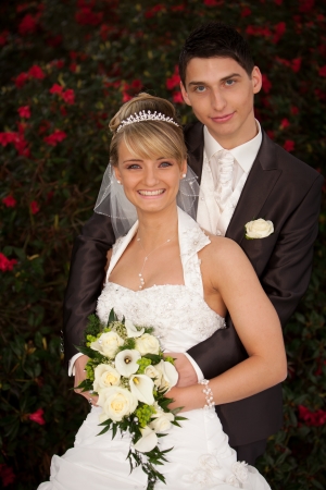 Bride leaning against groom  Young couple just married  groom and pretty bride are looking into the camera to the photograph other after the wedding ceremony  she is blond and wearing a nice diadem  background red roses and foreground yellow bouquet rosesの写真素材