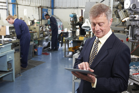 Owner Of Engineering Factory Using Digital Tablet With Staff In Background Owner Of Engineering Factory Using Digital Tablet With Staff In Background